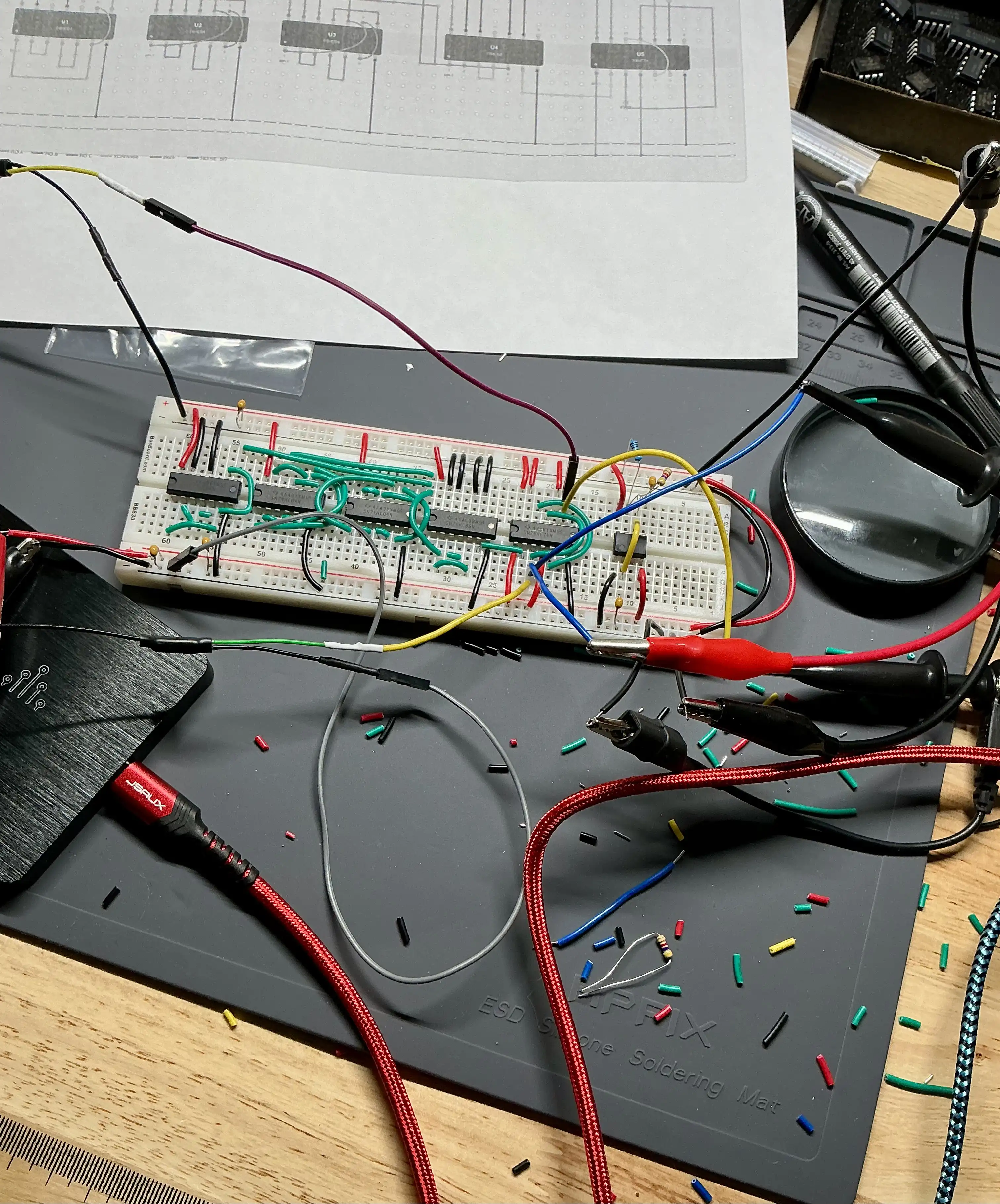 A breadboard full of 74-series logic chips and jumper wires on an ESD mat, with a printed schematic, scope probes, and a magnifier nearby