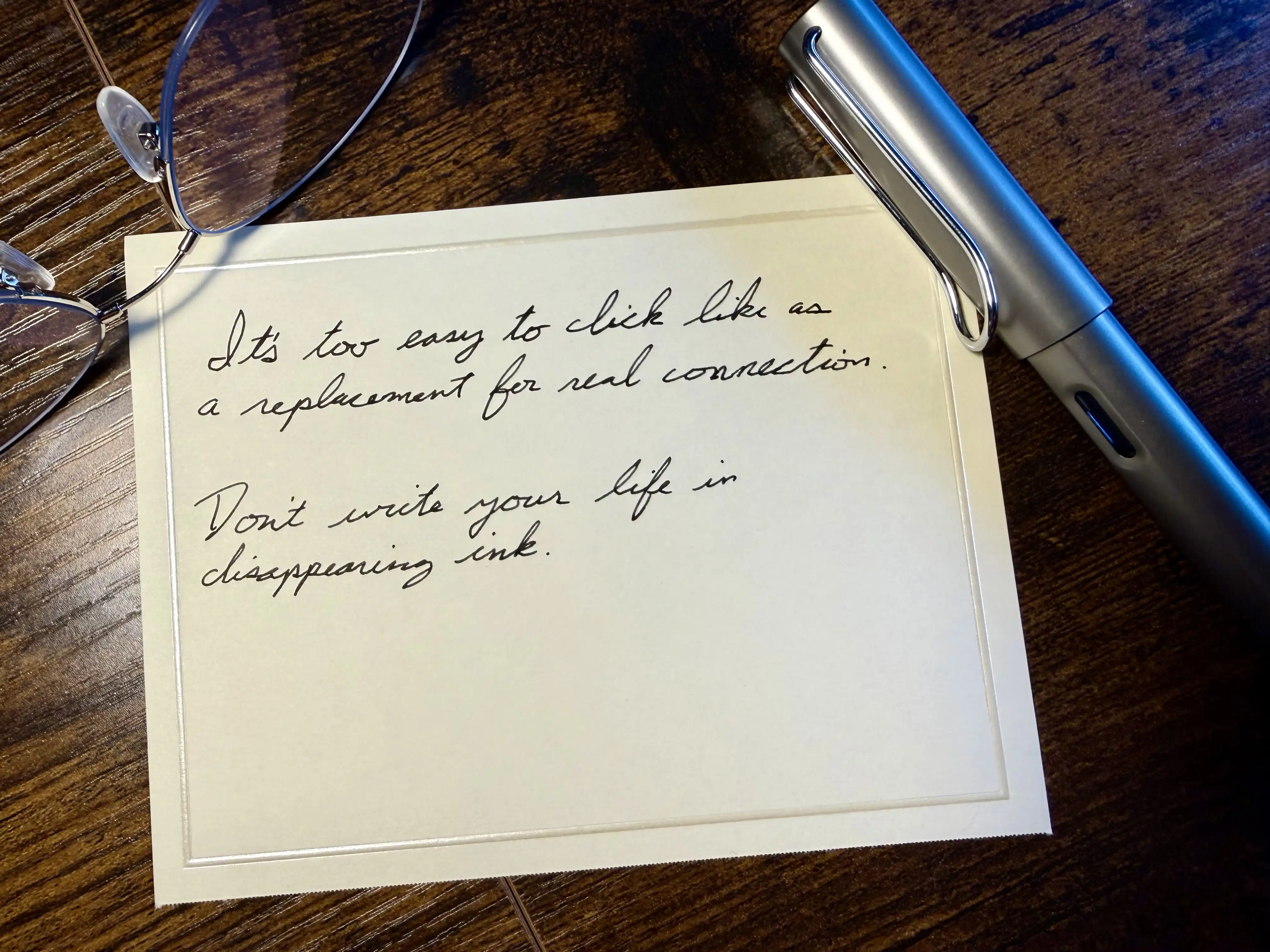 A handwritten note beside a fountain pen and reading glasses that reads: It’s too easy to click like as a replacement for real connection. Don’t write your life in disappearing ink.