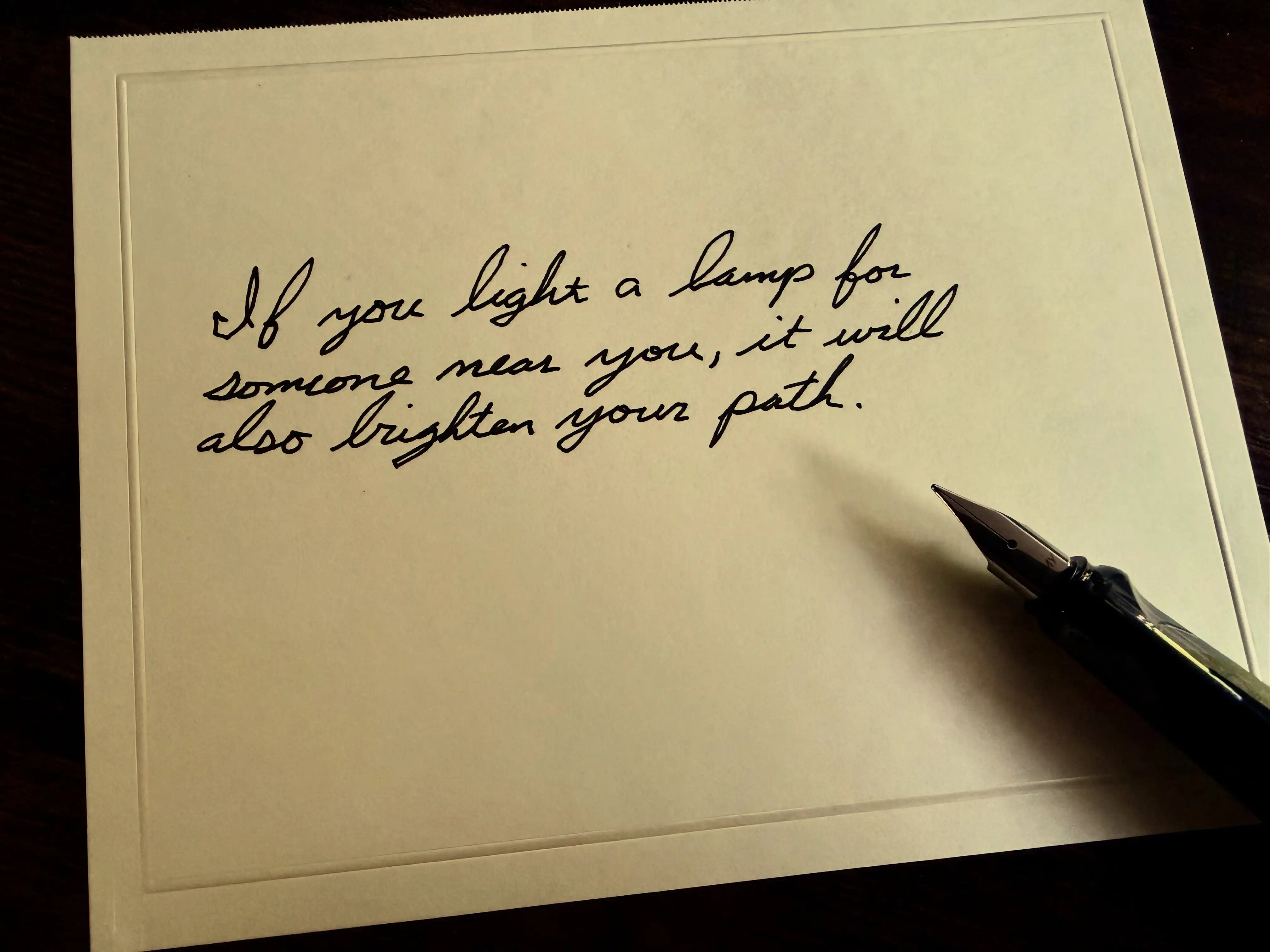A handwritten note beside a fountain pen that reads: If you light a lamp for someone near you, it will also brighten your path.