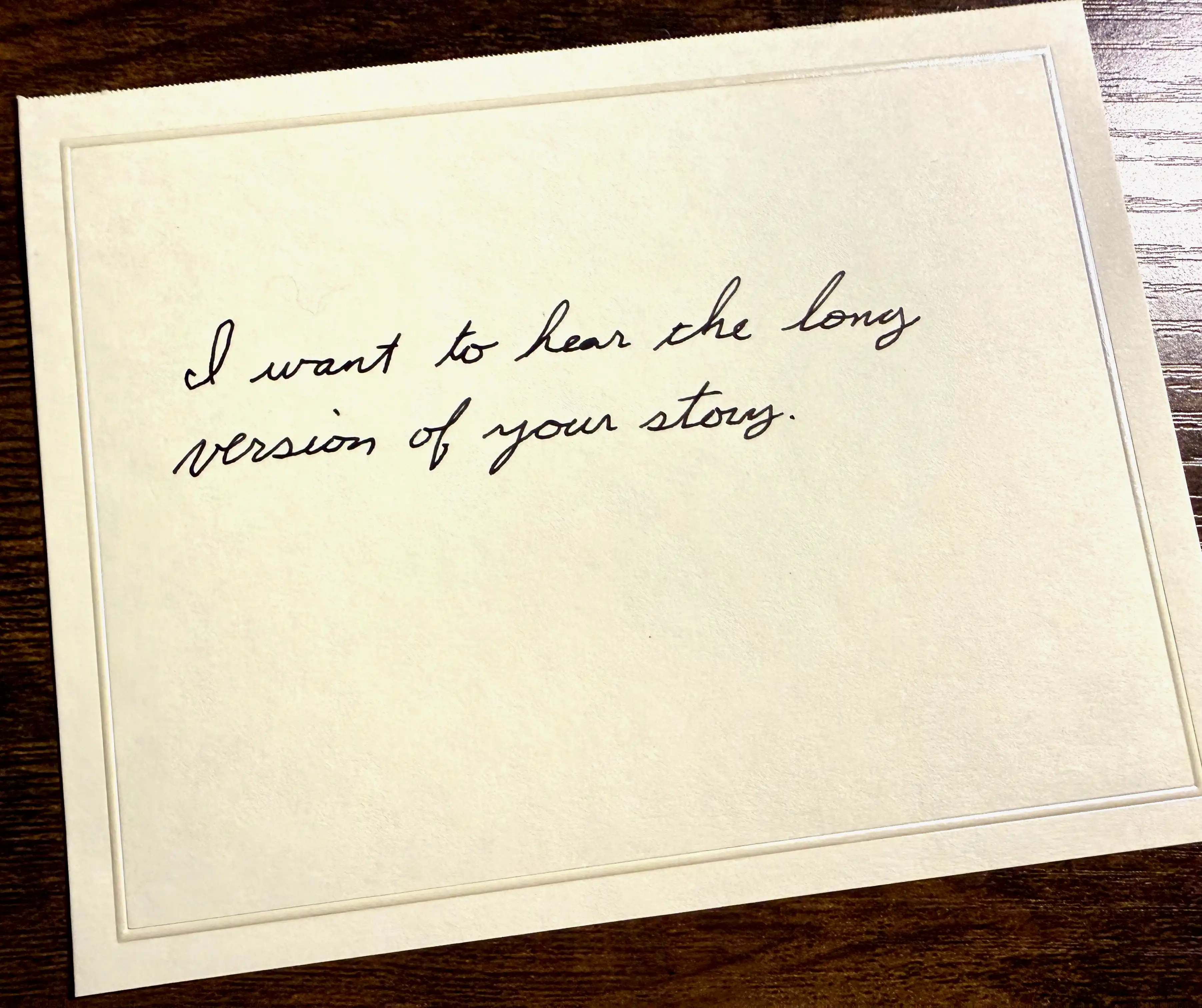 A handwritten note card on a wooden table that reads: I want to hear the long version of your story.