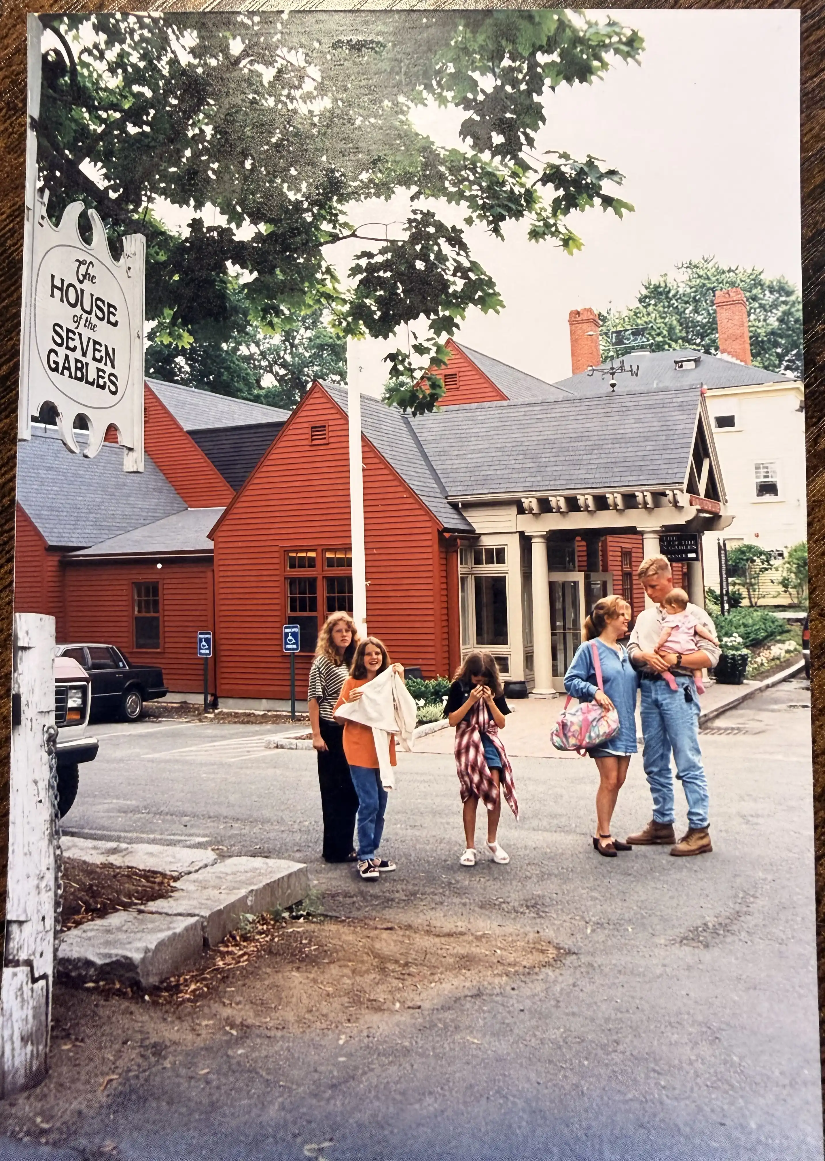 An old family photo outside the House of the Seven Gables in Salem, Massachusetts. A young man holds a baby girl in a pink outfit while a young woman smiles at them, and three girls stand nearby in front of the red clapboard buildings.