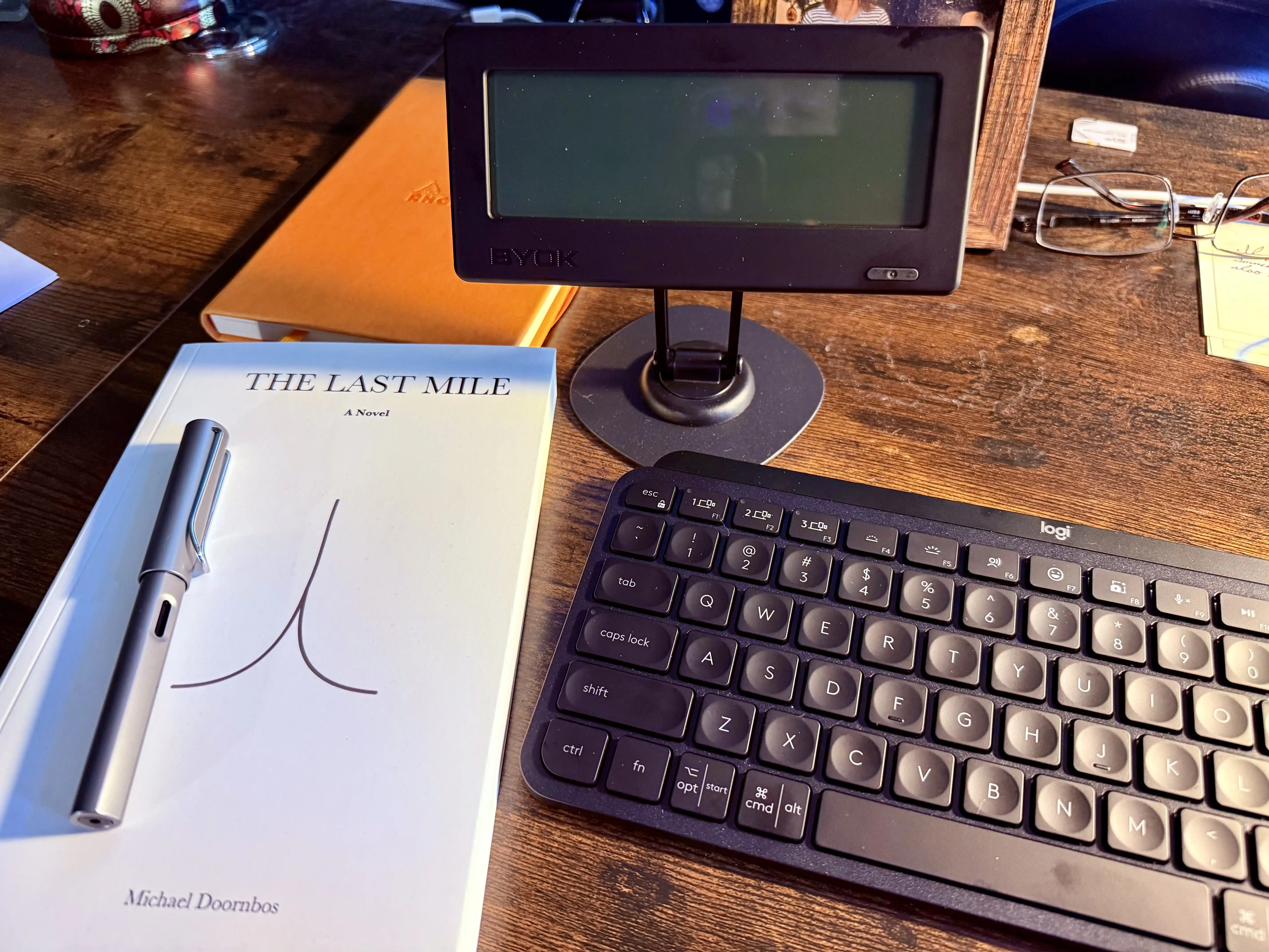 A bound copy of The Last Mile: A Novel by Michael Doornbos on a wooden desk with a fountain pen resting on the cover, next to a BYOK e-paper writing display, a Logitech keyboard, an orange Rhodia notebook, and a pair of glasses.